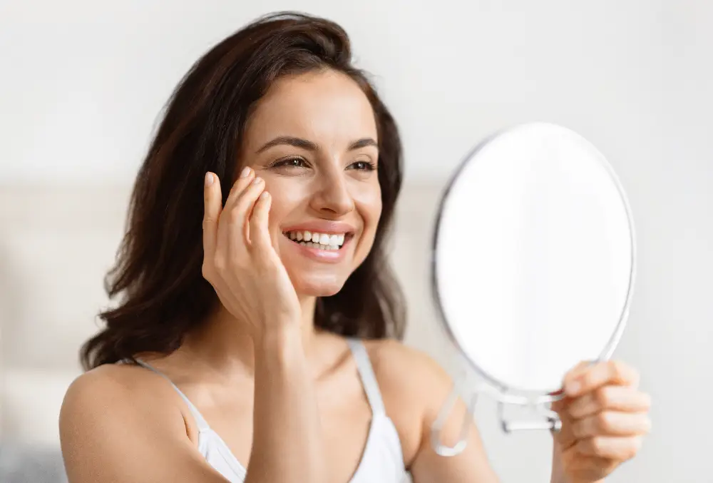 Woman smiling while looking in a mirror with radiant, healthy-looking skin.